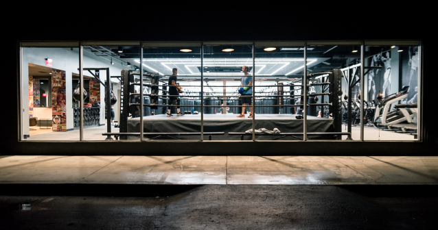 Interior view of a modern boxing gym at night, showing two athletes training inside a boxing ring surrounded by fitness equipment and punching bags.
